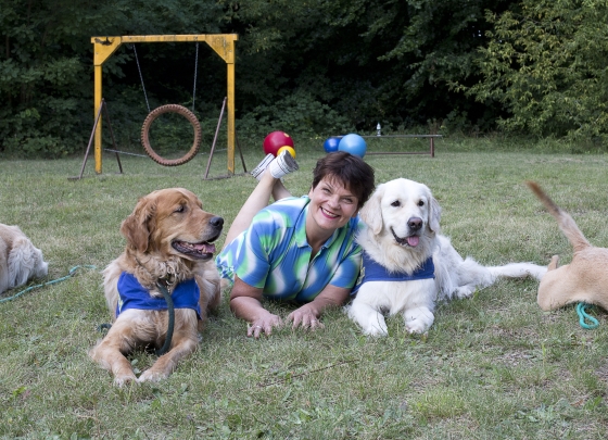 2015 Gayle Tufts mit Joschi und Mars 2015 Gayle Tufts mit Joschi und Mars. Das Foto zeigt eine Sommerwiese. Im Vordergrund liegt eine lachende Gayle Tufts bäuchlings auf der Wiese, die Beine baumeln in der Luft. Rechts neben Gayle liegt Mar,s weißer Golden Retrieve,r mit dem typisch entspannten Retrieverlächeln, links neben Gayle liegt Joschi, ein goldfarbener Golden Retriever, der interessiert zur Seite blickt. Beide Hunde tragen die blaue Kenndecke von HfH. Am rechten Bildrand ist noch ein Schwanz und ein Hundebein zu von einem, sich aus dem Staub machenden Hund, zu sehen
