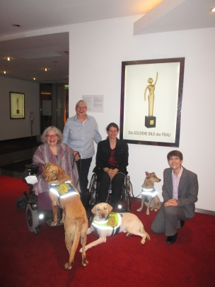 Auf dem Foto zeigt  das gutgelaunte HfH Team, dass zur Preisverleihung der Goldenen Bild der Frau nach Hamburg gefahren ist. Sabine H&auml;cker wird von den beiden Rollstuhlfahrerinnen Eileen und Jasmin und von Christel, die hinter den Rollst&uuml;hlen steht, begleitet. Sabine kniet auf dem Boden, neben ihr die beiden Assistenzhunde von Jasmin, sitzend Luna, mit blauer Kenndecke und vor ihr liegend, Snickers, ein heller Labrador mit gelber Kennedecke. Quincy, ein dunkel goldener Labrador, ist der Assistenzhund von Eileen. Er steht mit seinen Vorderpfoten auf dem Rollstuhl von Eileen und schaut interessiert in die Kamera. Snickers hat seinen Blick auch auf den Fotografen gerichtet.