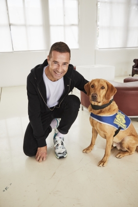 Ein Kai Pflaume kniet mit einem Bein, neben ihm sitzt Quincy, ein dunkel goldfarbener Labrador R&uuml;de. Quincy tr&auml;gt die blaue Kenndecke von Hunde f&uuml;r Handicaps. Die Kenndecke kennzeichnet ihn als gepr&uuml;ften Assistenzhund. Das Foto wurde f&uuml;r die Vorstellung der Goldene Bild der Frau Preistr&auml;gerinnen 2020 im Studio gemacht.