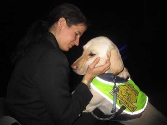 Das Foto zeigt Jasmin, wie sie liebevol mit ihrer Hand ihren hellen Labradorr&uuml;den Snickers am Kopf krault. Snickers hat seine beiden Vorderpfoten auf den Scho&szlig; von Jasmin gelegt und genie&szlig;t sichtlich die Streicheleinheiten. Snickers tr&auml;gt die gelbe Kenndecke von HfH, die ihn als Assistenzhund in Ausbildung ausweist.