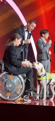Ein Bild von der B&uuml;hne bei der Preisverleihung der Goldenen Bild der Frau. Im Vordergrund steht Jasmin mit Rollstuhl, vor ihr steht Snickers. Snickers ist ein heller Labradorr&uuml;de, der Assistenzhund in Ausbildung von Jasmin. Er tr&auml;gt die gelbe Kenndecke von HfH. Jasmin h&auml;lt eine Tupperdose in der Hand, die von Snickers mit seinen Z&auml;hnen ge&ouml;ffnet wird. Hinter Jasmin steht der Moderator Kai Pflaume, der ein Mikrophon in der Hand h&auml;lt und den Auftritt des Teams moderiert. Sabine H&auml;cker steht hinter Kai Pflaume, sie hat ebenfalls ein Mikrophon in der Hand und erkl&auml;rt die Arbeit von Hunde f&uuml;r Handicaps