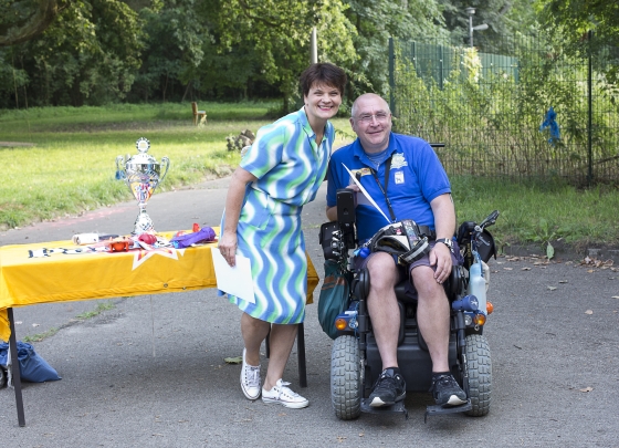 2015 Gayle Tufts und Jens Schmidt 2015 Gayle Tufts und Jens Schmidt. Ein Sommerbild. Das Foto zeigt Gayle Tufts im Sommerkleid. An ihrer Seite ein freudiger Jens Schmidt mit seinem Rollstuhl .Jens trägt das blaue HfH Shirt. Neben den beiden steht ein Tisch mit gelber Tischdecke und Aufschrift Predigee. Auf dem Tisch steht der silberne Wanderpokal von HfH.