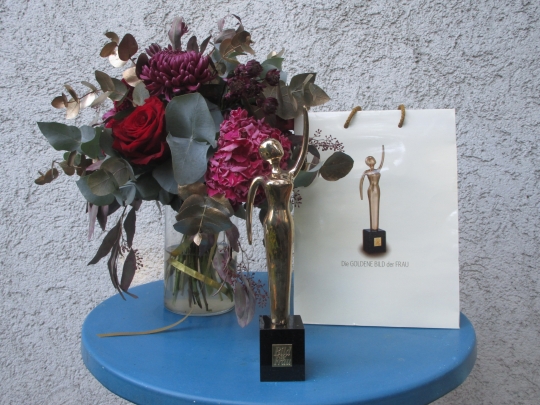 Das Foto zeigt die Preise, die Sabine H&auml;cker bei der Verleihung der Goldenen Bild der Frau bekommen hat. Auf einem kleinen blauen Tisch steht die Troph&auml;e "Goldene Bild der Frau". Der Preis ist eine kleine goldene Statur, die auf einem schwarzen Sockel steht. Es stellt eine stilisierte, elegante Frau dar, die stolz einen Arm in die H&ouml;he reckt. Daneben steht eine Vase mit einem Blumenstrau&szlig; und deine Urkunde