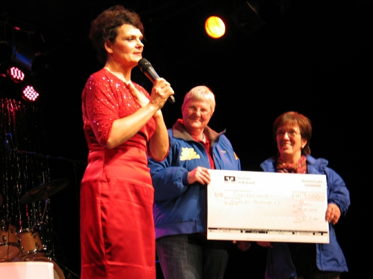2011 Gayle Tufts 2011 Scheckübergabe von Gayle Tufts an HfH. Gayle Tufts steht in einem roten, schicken Kleid auf der Bühne, in der Hand hält sie ein Mikrofon. Ein überdimensionaler Scheck wird an HfH überreicht.