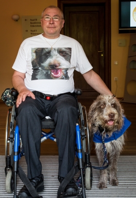 Rieke und Jens Rieke und Jens, ein perfektes Team auf diesem Foto. Rieke, die grau-braun-weiße Mischlingshündin steht neben dem Rollstuhl von Jens. Farblich sind beide perfekt auf einander abstimmt: blauer Rollstuhl passend zur blauen Kenndecke von HfH, die Rieke trägt. Jens hat seine Hand zum Knuddeln auf Riekes Rücken gelegt. Während Jens direkt in die Kamera blickt, schaut Rieke amüsiert in die andere Richtung. Ihr Blick zeigt, irgendetwas scheint sie zu erheitern? Was es wohl ist? Riekes größter Fan ist natürlich Jens selbst, denn er trägt ein weißes T-Shirt mit einem großen Foto von Riekes Kopf.