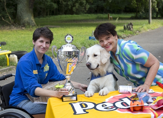 2015 Gayle Tufts und Jasmin Stoiber 2015 Gayle Tufts und Jasmin Stoiber. Eine stolze Jasmin sitz draußent im Rollstuhl an einem Tisch. Der Tisch ist mit einer Tischdecke von Pedigree bedeckt. Vor ihr steht ein großer, silberner Pokal. Die Aufschrift am Pokal lautet: Wanderpokal, Hunde für Handicaps, Behinderten-Begleithunde. Mars, ein weißer Golden Retriever, steht auf zwei Beinen und hat seine Vorderpfoten auf den Tisch gelegt.Mars trägt die blaue Kenndecke von HfH. Daneben beugt sich lächelnd Gayle Tufts über den Hund. Jasmin hat eine kleine Kiste mit einer Schleife auf dem Schoß.