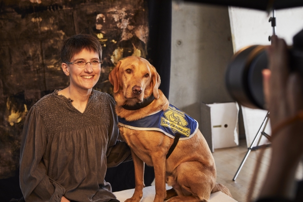 Sabine Häcker und Quincy Das Foto zeigt Sabine Häcker beim Fotoshooting für die Goldene Bild der Frau. Sabine sitzt auf einer weißen Kiste, neben ihr sitzt Quincy, der die blaue Kenndecke von HfH trägt. Sabine lächelt in die Kamera, während Quincy interessiert in Richtung Kamera blickt.