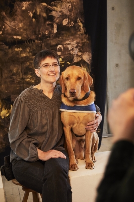 Bei dem offiziellen Fotoshooting bei der Goldenen Bild der Frau sitzt Sabine H&auml;cker auf einem Hocker, neben ihr sitzt der dunkel goldene Labradorr&uuml;de Quinncy auf einer wei&szlig;en Kiste. Sabine l&auml;chelt in die Kamera und hat einen Arm um Quincy gelegt. Quincy schaut an der Kamera vorbei und hat seinen Blick jemandem interessanteren zugewandt. Er tr&auml;gt die blaue Kenndecke von HfH