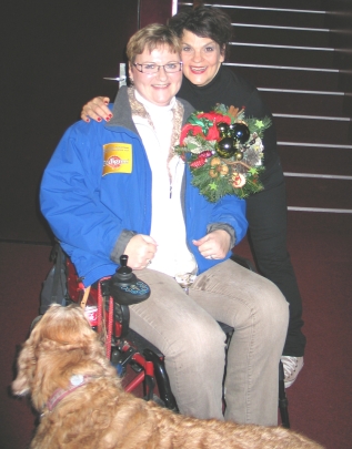 2011 Ursula Heiner und Gayle Tufts 2011 Ursula Heiner und Gayl Tufts. Auf dem Foto sieht man Ursula Heiner im Rollstuhl. Sie trägt die blaue HfH Jacke. Neben ihr beugt sich Gayle Tufts zu ihr hin und hat einen Arm um Ursula gelegt, in der anderen Hand hält Gayle einen Blumenstrauß. Vor dem Rollstuhl steht ein dunkler Golden Retriever.