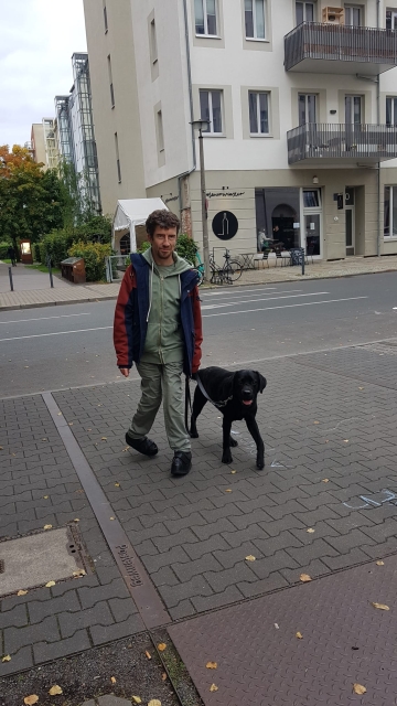 Christian und Toby Mann (Christian) mit schwarzem Labrador Retriever (Toby) an der Leine laufen auf den Bürgersteig. Beide schauen in die Kamera. Toby geht fuß.