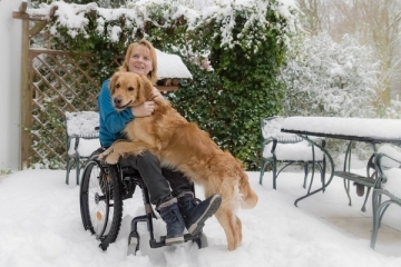 Pluto & Angela Ein Winterfoto!. Angela steht mit ihrem Rollstuhl auf der dick verschneiten Terrasse im Garten. Tisch, Stühle und die Pflanzen haben dicke Schneemützen auf. Pluto, der goldene Golden Retriever steht neben dem Rollstuhl und hat seine Vorderpfoten auf den Schoß von Angela gelegt. Angela hat ihre Arme um Plutos Hals in das wuschelig, warme Fell gelegt. Das ist Retriever Wetter, Pluto sieht so aus, als ob er sich gleich in den Schnee stürzen wird, einmal wälzen oder zumindest einmal mit der Nase durch den Schnee wühlen.