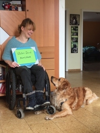 Pluto und Angela Dieses Foto zeigt Pluto, ein goldener Golden Retriever und Angela in ihrem Rollstuhl. Angela hat ein grünes Schild in ihren Händen. Auf dem Schild steht in großer Schreibschrift "Vielen Dank Pluto und Angela". Die freudestrahlende Angela schaut zu Pluto, der zu ihren Füßen auf dem Boden Platz gemacht hat. Pluto schaut neugierig zu Angela. Da möchte man doch gerne wissen, was Angela ihm versprochen hat.