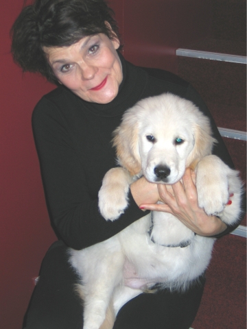2011 Gayle Tufts Das Foto zeigt eine sitzende Gayle Tufts, die einen sehr hellen Golden Retriever Welpen im Arm hält. Gayle Tufts hat ein schwarzes Oberteil und eine schwarze Hose an. Der kleine Welpe hat seine Pfoten über den Arm von Gayle Tufts gelegt, wärend er auf dem Schoß sitzt. Das kleine dicke Bäuchlein des Welpen, drängt sich dem Betrachter schmunzelnd entgegen.