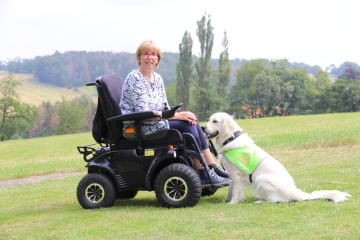 Ellis und Anke Ellis und Anke. Anke steht mit ihrem Elektrorollstuhl auf einer Wiese. Vor ihr sitzt die helle Golden Retrieverhündin Ellis. Sie trägt eine gelbe Kenndecke von HfH.