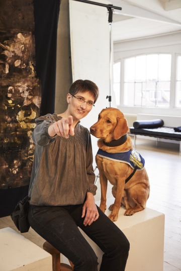 Sabine Häcker, Vereinsvorsitzende von Hunde für Handicaps e.V., mit Assistenzhund Quincy Sabine und Quincy beim Fotoshooting zur Vorstellung der Preisträgerinnen der Goldenen Bild der Frau. Sabine und Quincy sitzen auf einer weißen Kiste in einem Fotostudio. Sabine hat ihren rechten Arm ausgestreckt und zeigt mit ihrem Finger in Richtung Kamera. Quincy schaut erwartungsvoll auf Sabines Arm. Quincy trägt die blaue Kenndecke von HfH